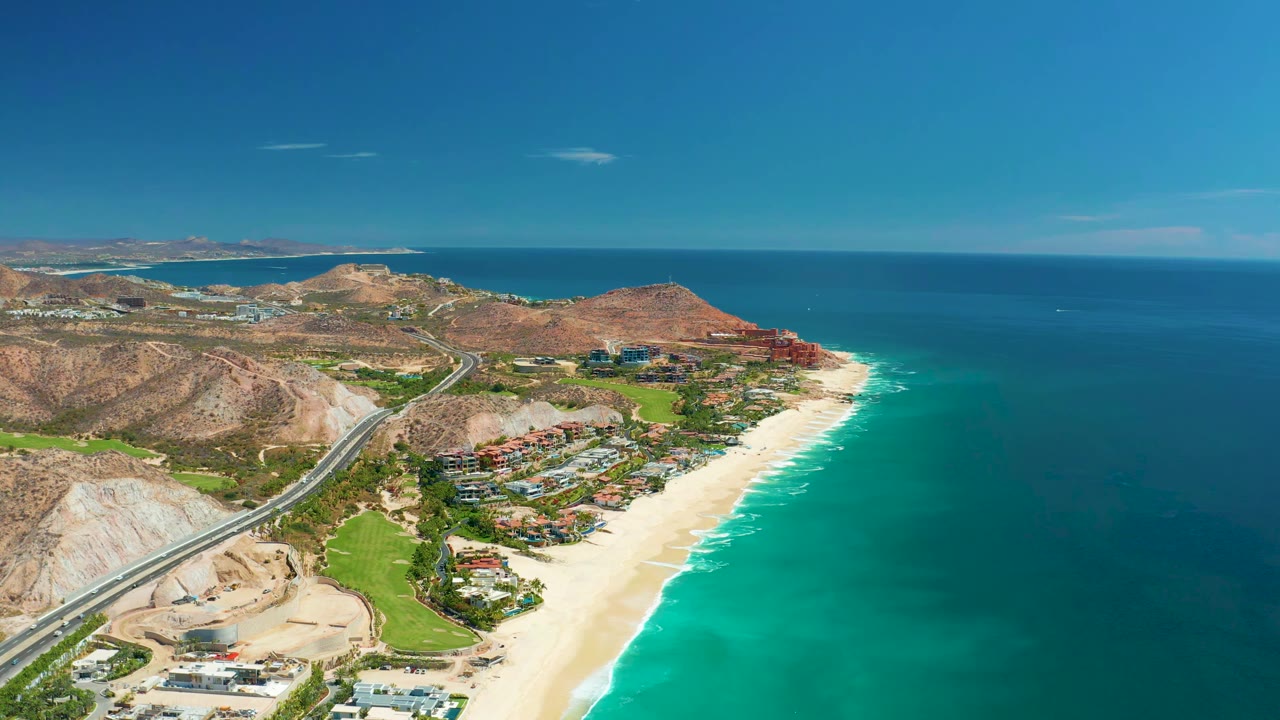 Wide aerial view at 0:01 over Marquis Los Cabos, Mexico, with the road cutting through dry hills toward beachside buildings and clear blue ocean, with no visible people.