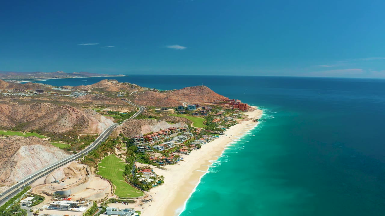 Aerial frame from Marquis Los Cabos, Mexico, showing white surf along the sandy shore, clustered coastal buildings, dry hills, a road, and open ocean, with no visible people.