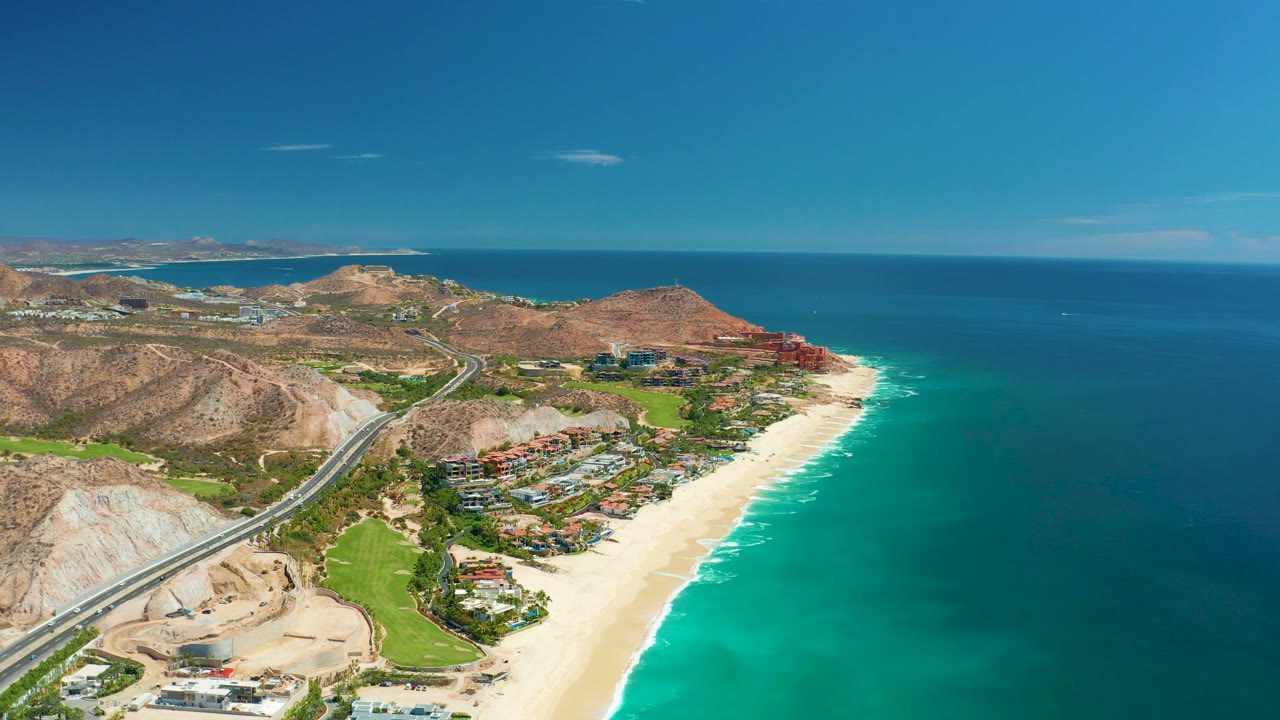 At 0:03, the aerial view over Marquis Los Cabos, Mexico, shows construction earthwork beside the road, green landscaped patches, the beach, and a broad blue ocean, with no visible people.
