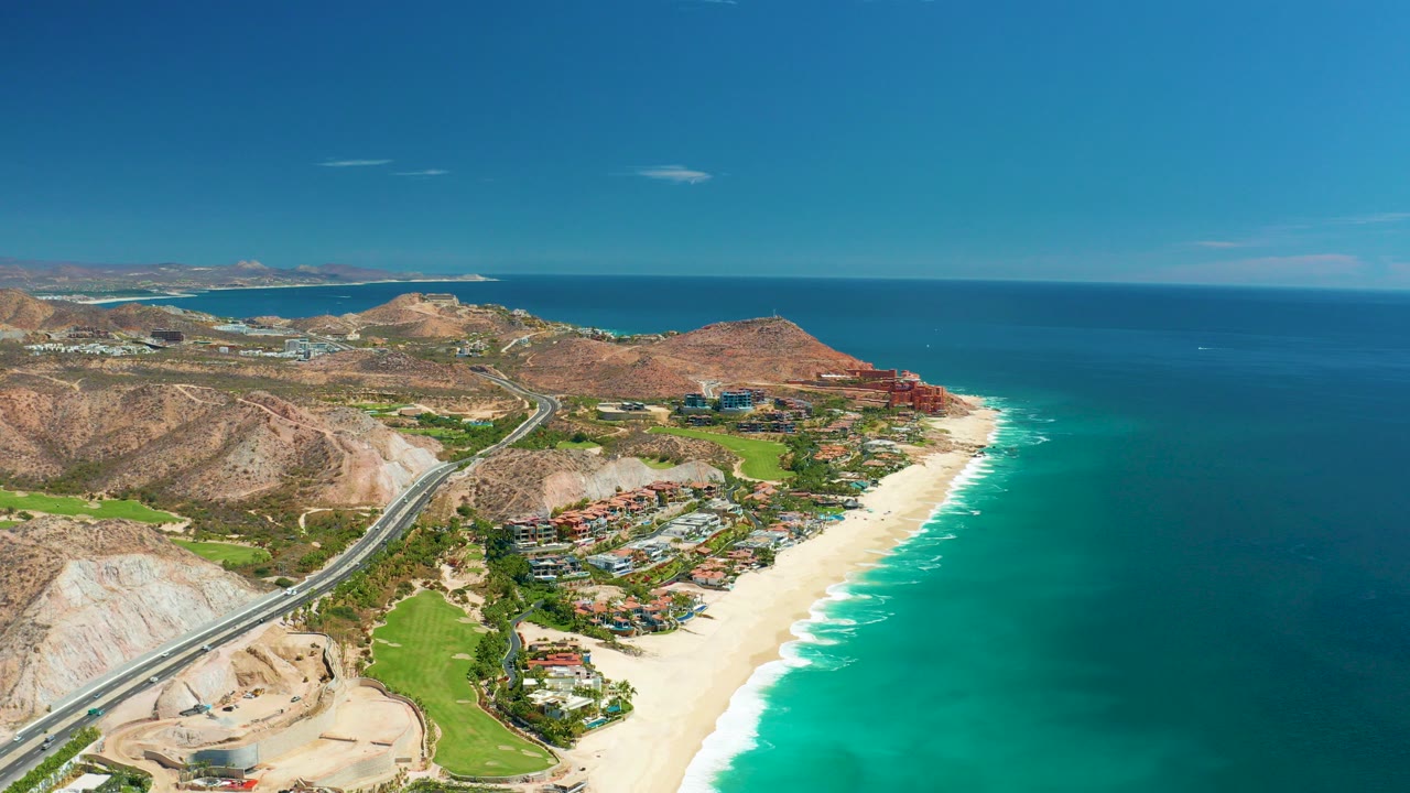 A high aerial frame over Marquis Los Cabos shows the coastal highway at left, sandy beach at lower right, turquoise ocean beyond, low mountains, developed shoreline, and no visible people.