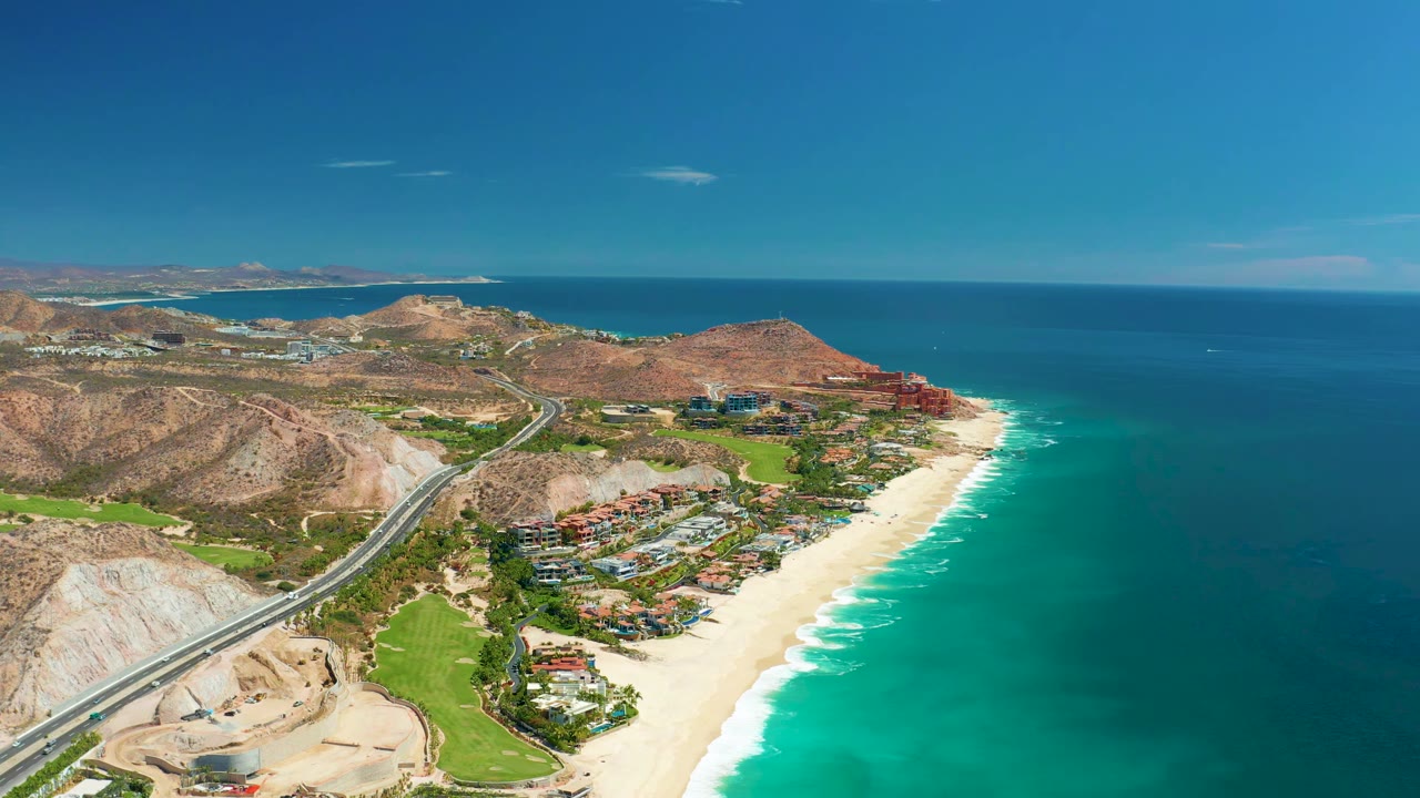 At 0:01, the Marquis Los Cabos coastline remains in a wide aerial composition with bright sand, blue-green water, hillside terrain, a curving road, clustered buildings, and no visible people.