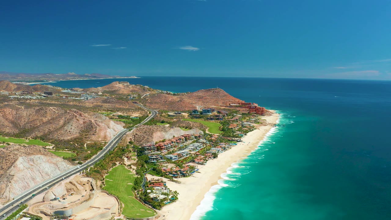 At 0:02, the aerial view over Marquis Los Cabos shows the surf line brighter along the beach, the coastal road cutting through the hills, ocean filling the right side, and no visible people.