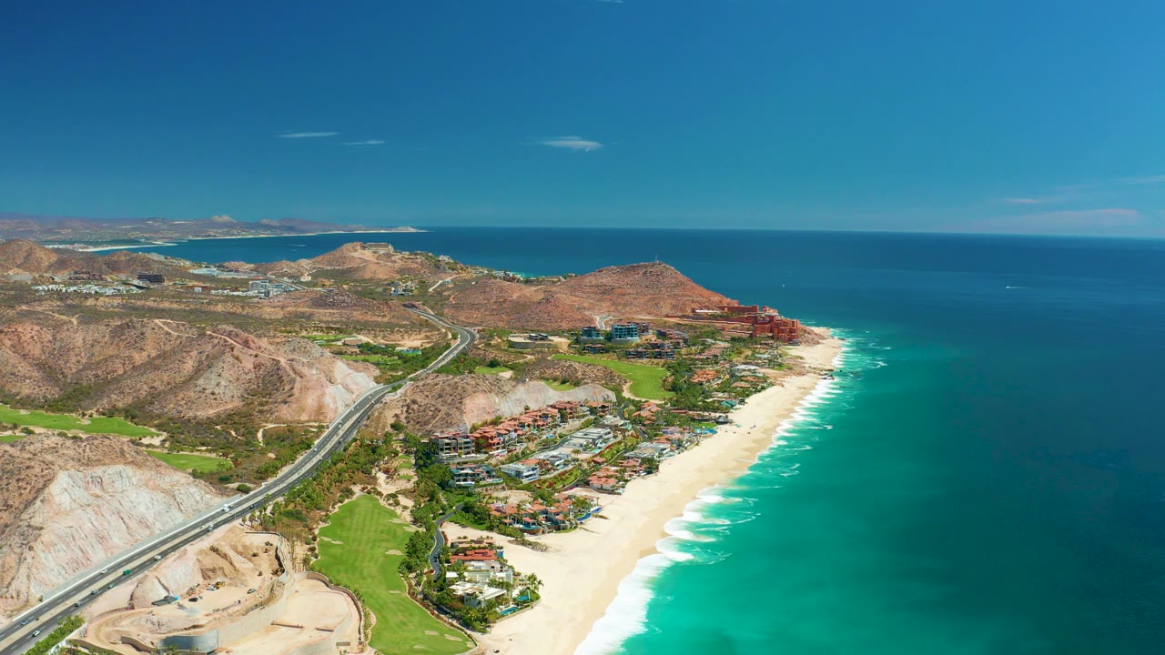 Aerial coastline with highway, beach surf, ocean horizon, hills, and buildings.