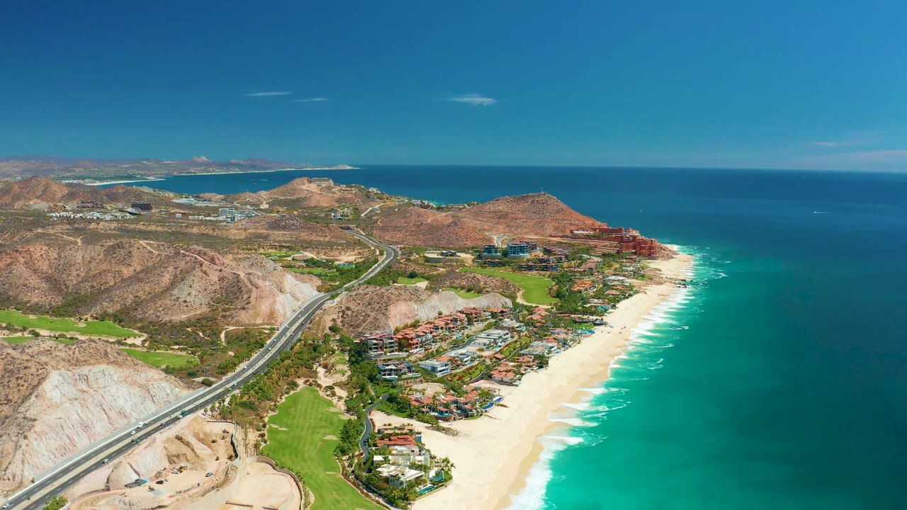 Aerial view in Marquis Los Cabos with the road running from the lower left toward hillside buildings and a bright sandy shoreline, no visible people.