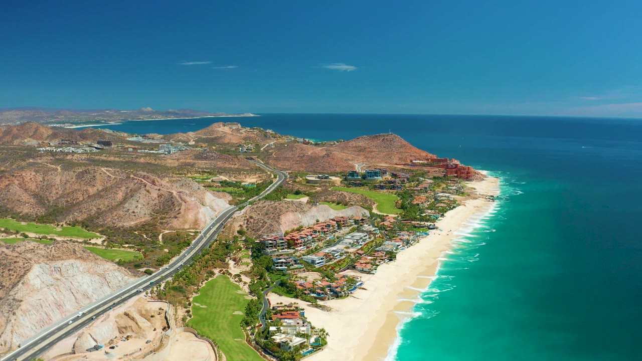 Aerial coastline with highway, beach surf, ocean, and arid hills.