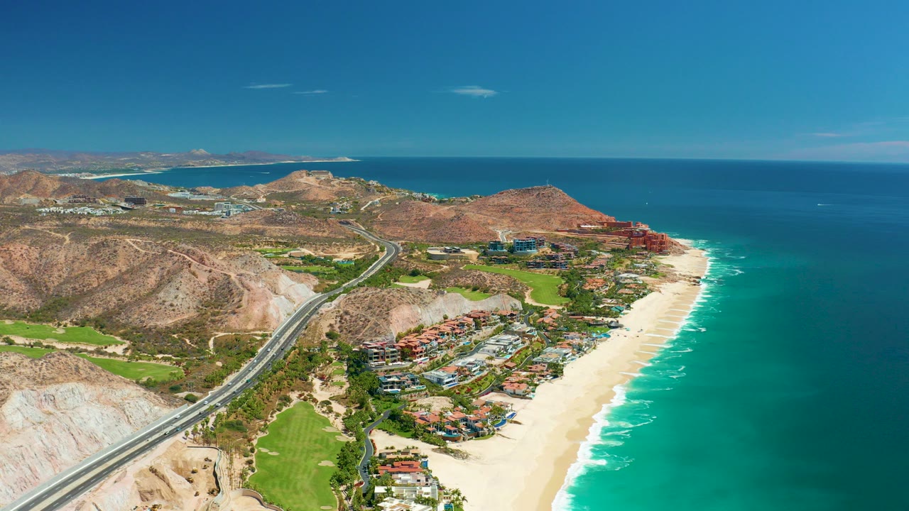 Aerial view over Marquis Los Cabos, Mexico, with a coastal highway at left, green landscaped areas below, sandy beach and turquoise ocean at right, and no visible people.