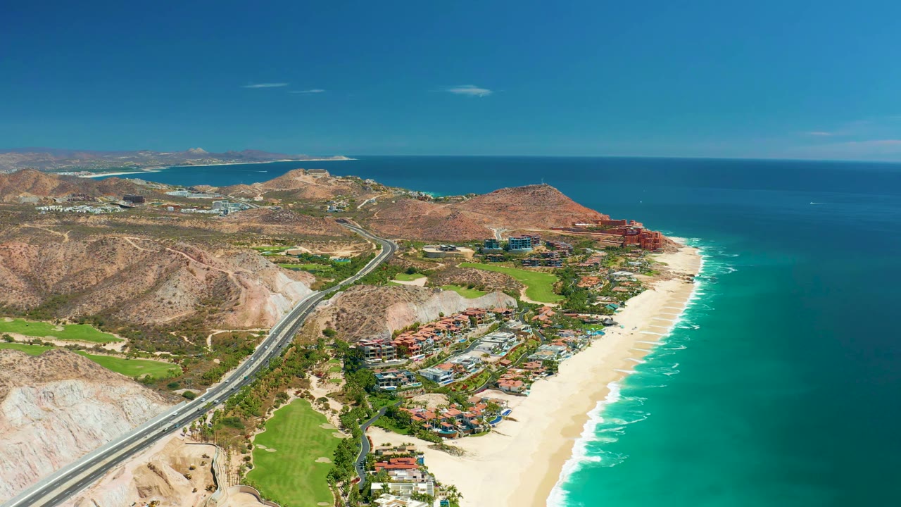 High aerial frame in Marquis Los Cabos, Mexico, showing the highway curving past hills and green grounds while the sandy beach and ocean fill the right side, with no visible people.