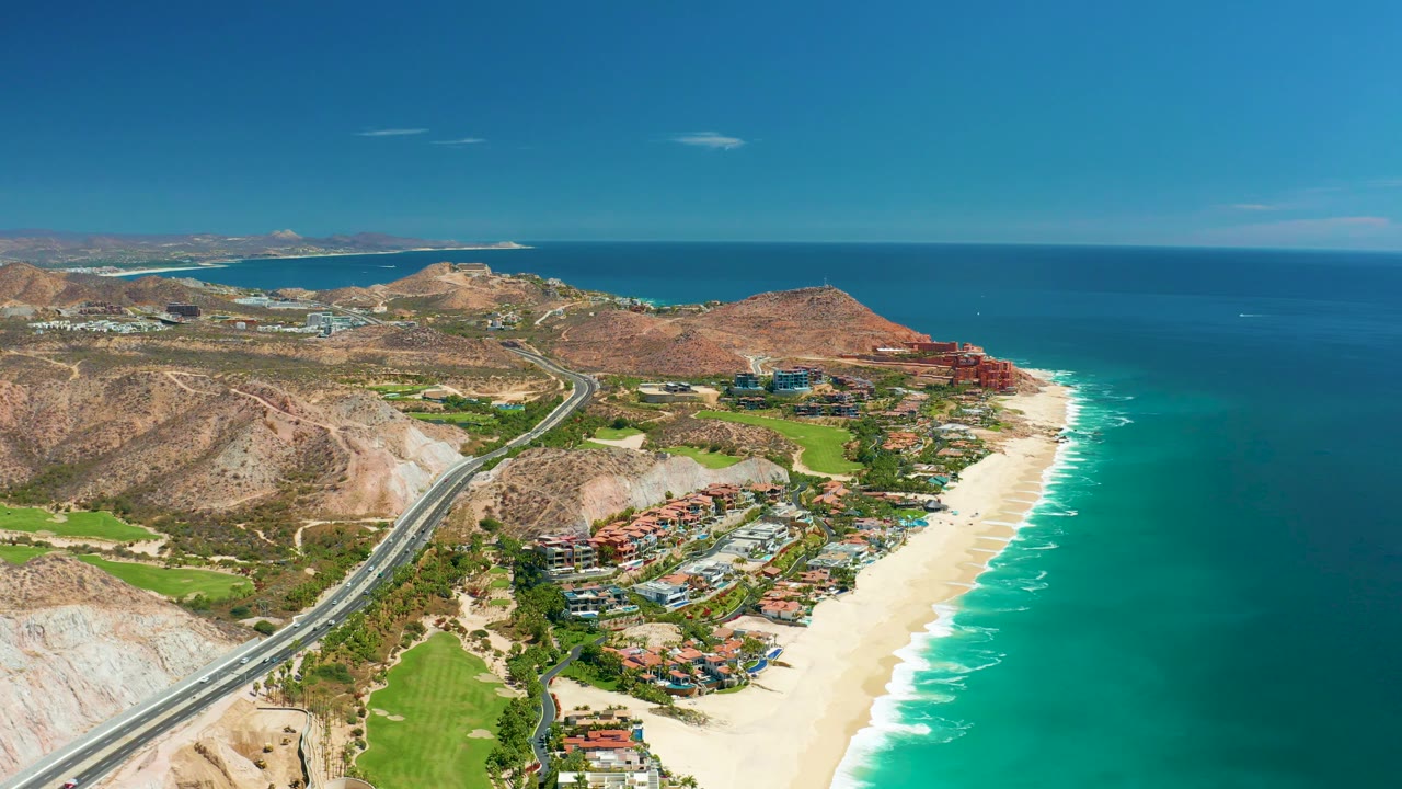 Aerial coastline with highway, green fairway-like areas, beach, and ocean surf.