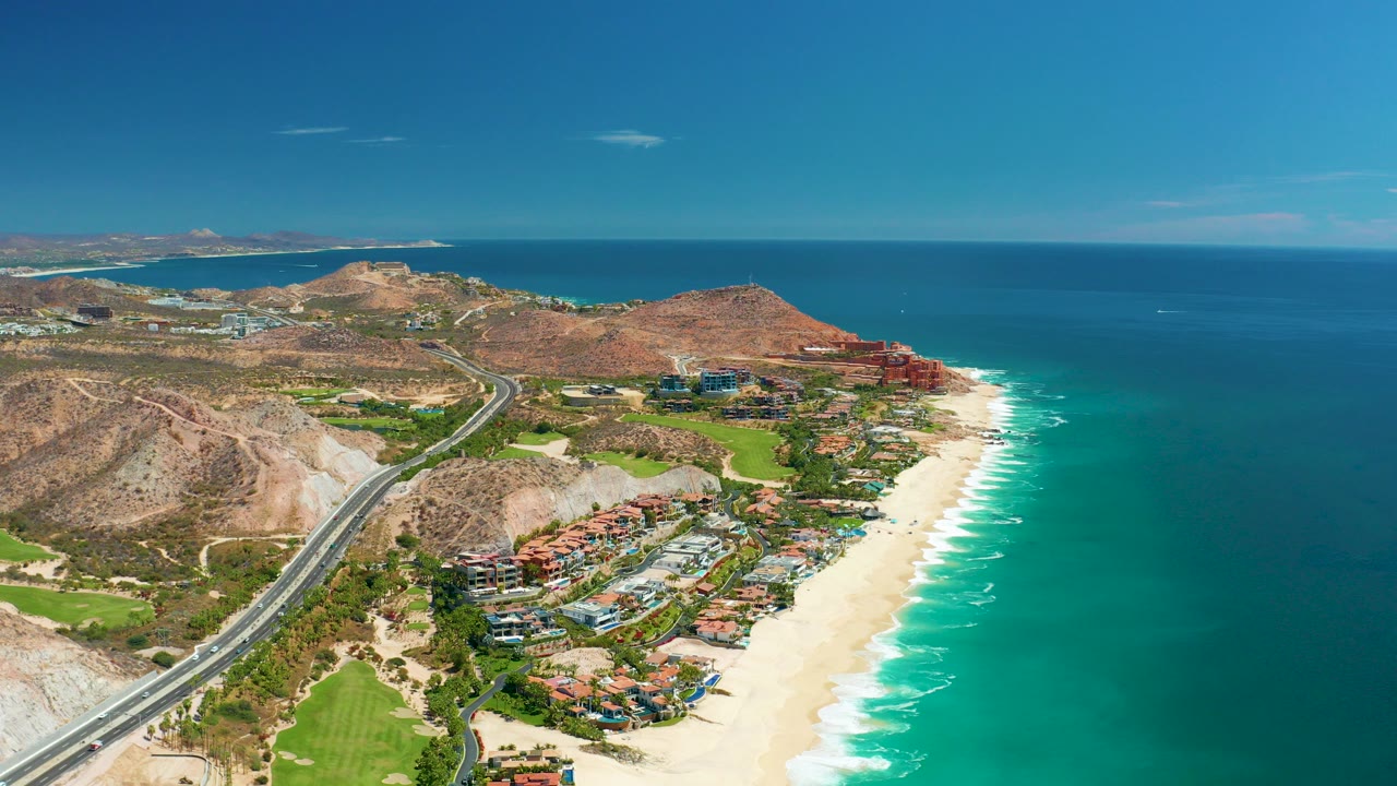 Opening aerial frame over Marquis Los Cabos, Mexico shows a coastal highway at left, sandy beach and turquoise surf at right, rocky hills and buildings inland, and no visible people.