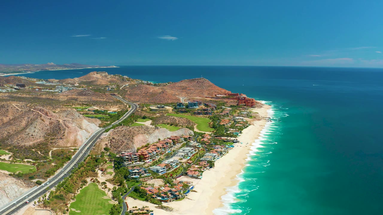 At 0:02, the Marquis Los Cabos coastline remains in a wide drone composition, with the curving road leading past rocky hills toward beach buildings and the blue ocean, with no visible people.