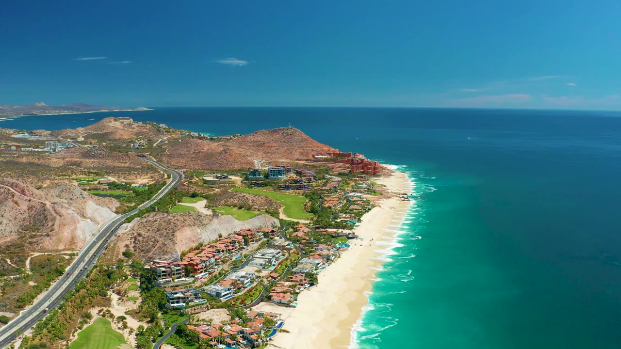 Aerial view in Marquis Los Cabos, Mexico with the highway curving through dry hills, clustered coastal buildings below, white sand and surf at right, and no visible people.