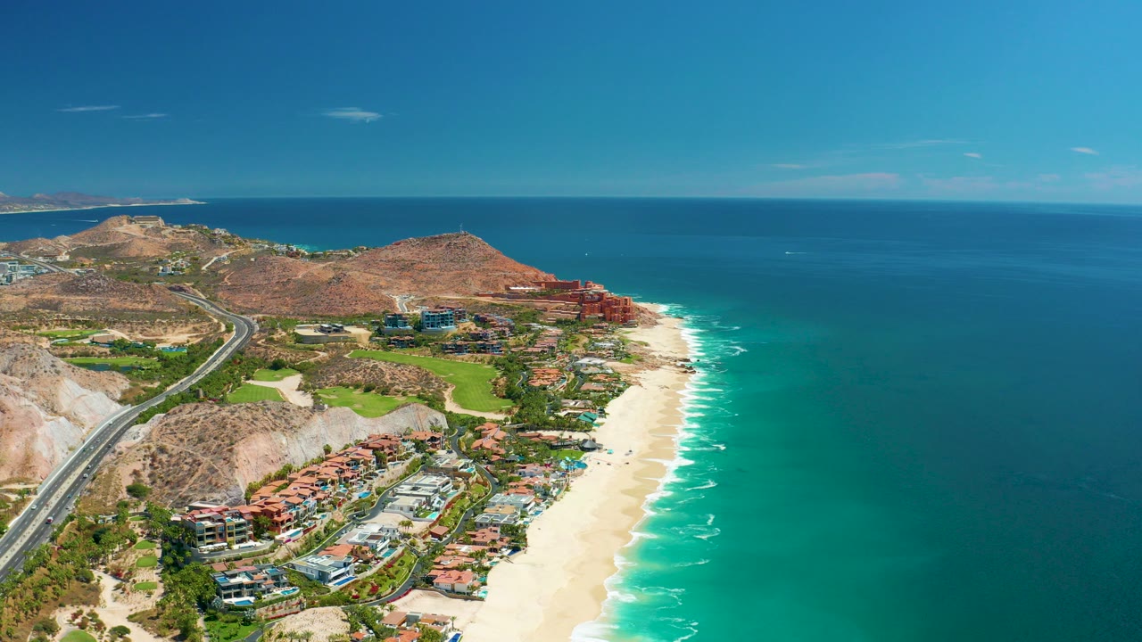 Opening aerial frame over Marquis Los Cabos, Mexico, with a coastal highway on the left, hillside homes below, a sandy beach and surf line through the center, turquoise ocean to the right, and no visible people.