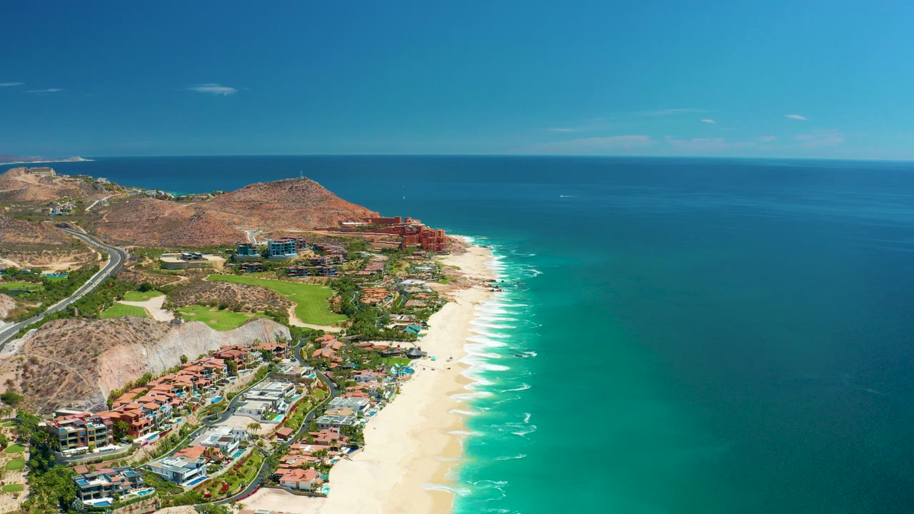 Opening aerial frame over Marquis Los Cabos, Mexico, showing pale beach curving beside turquoise water, coastal buildings, a road, dry hills, and a clear horizon.