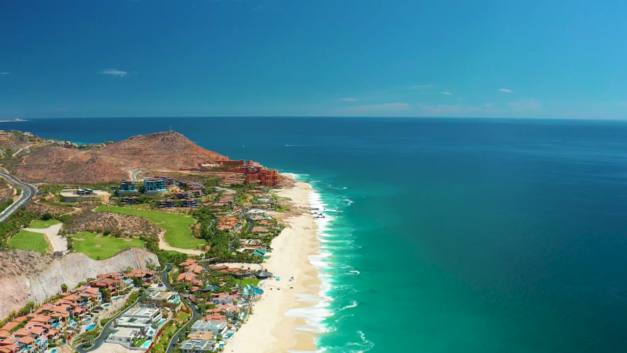 Aerial frame over Marquis Los Cabos, Mexico showing red-roofed resort buildings, a sandy shoreline, turquoise ocean water, dry hills, a road at left, and no visible people.
