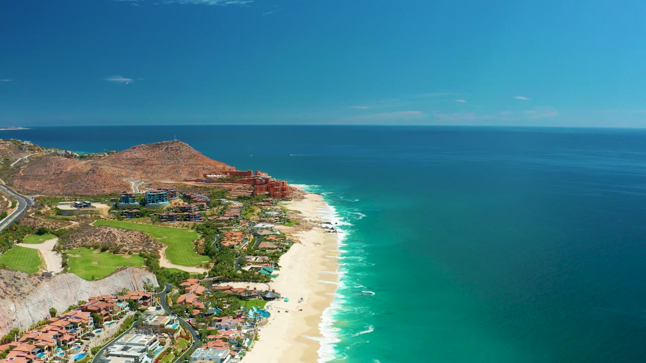 Opening aerial frame over Marquis Los Cabos, Mexico, showing a sandy beach beside turquoise water, coastal buildings, a road at far left, a brown hill, and no visible people.