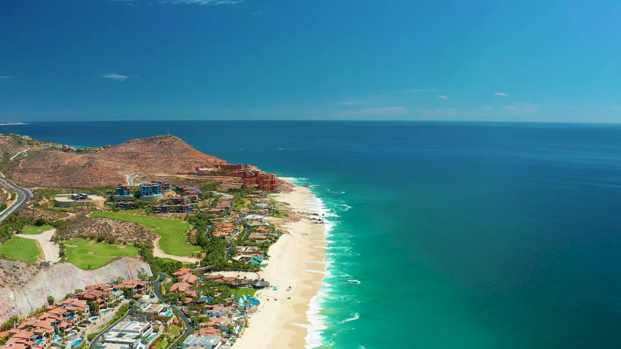 Aerial view at 0:01 over Marquis Los Cabos, Mexico, with the beach and bright surf running diagonally past buildings and green areas, no visible people.
