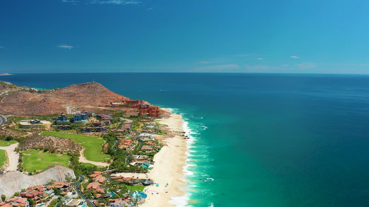 Wide aerial frame over Marquis Los Cabos, Mexico showing resort buildings and golf course beside a sandy beach, turquoise ocean, brown hills, blue sky, and no visible people.