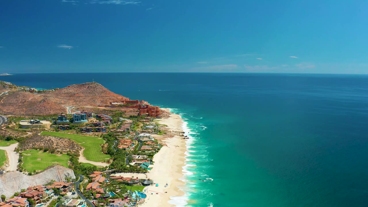 Aerial view of Marquis Los Cabos with the beach running along the right edge of the resort grounds, ocean stretching to the horizon, hillside behind the buildings, and no visible people.