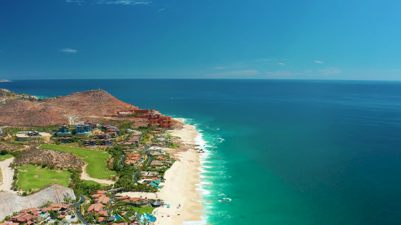 Aerial coastline with golf course, resort roofs, sandy beach, and ocean.