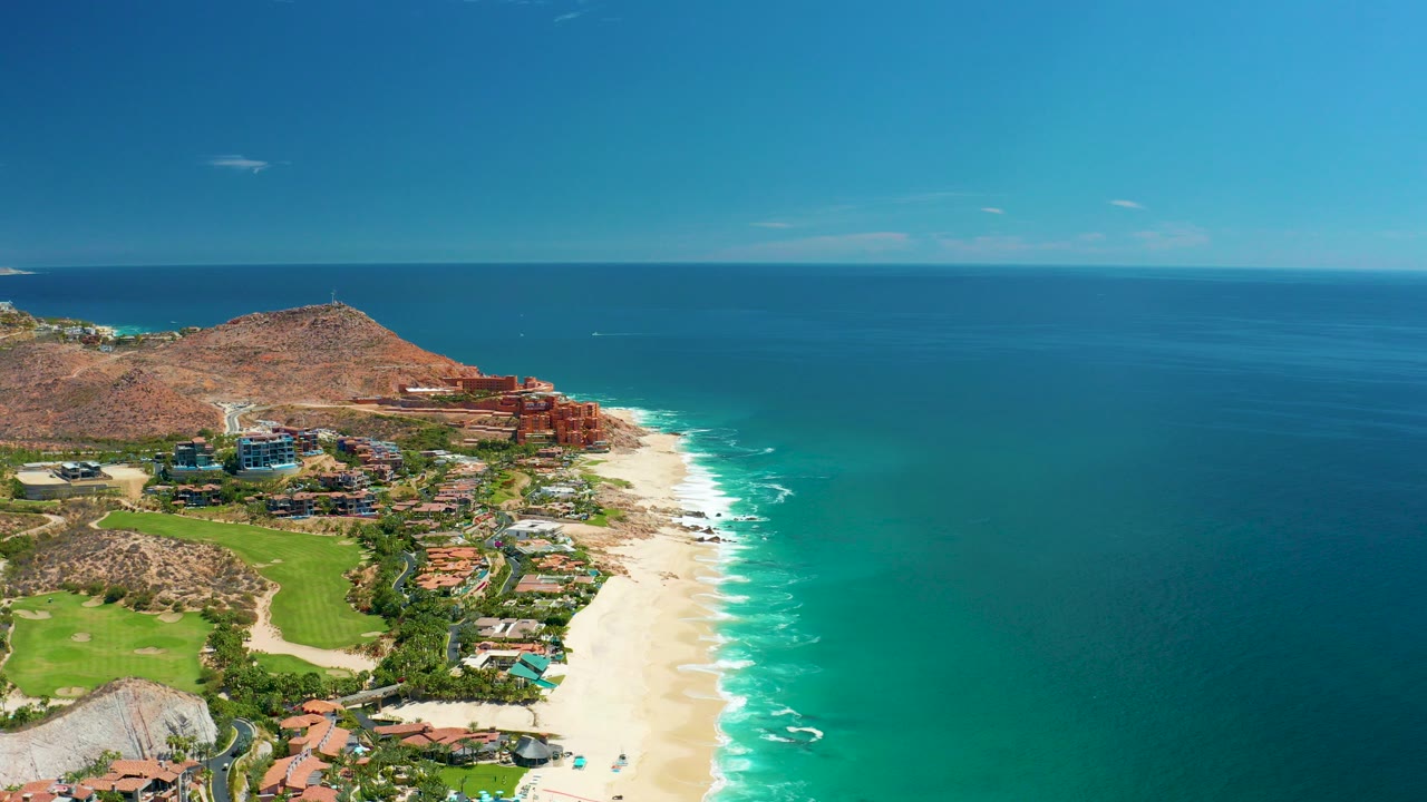 Aerial frame over Marquis Los Cabos, Mexico, showing pale beach, turquoise surf, coastal buildings, green space, rocky hill, clear sky, and no visible people.