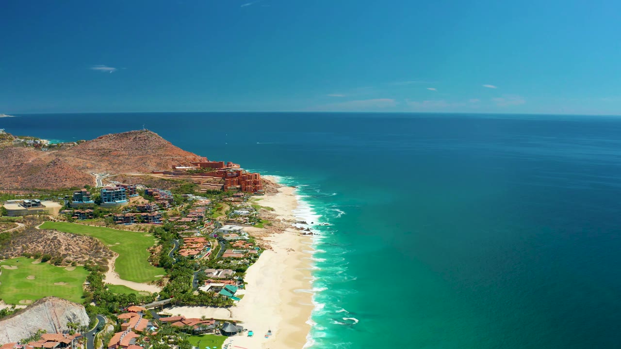 Aerial coastline frame from Marquis Los Cabos showing clustered buildings near a rocky hill, pale sand, turquoise water bands, white surf lines, and no visible people.