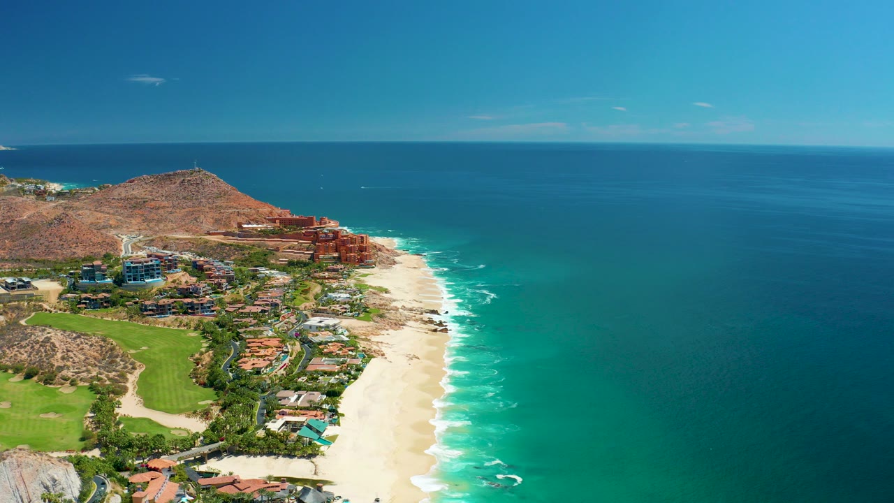 Aerial frame at Marquis Los Cabos BCS Mexico showing a sandy beach beside turquoise water, hillside buildings, reddish rock, green grounds, clear sky, and no visible people.