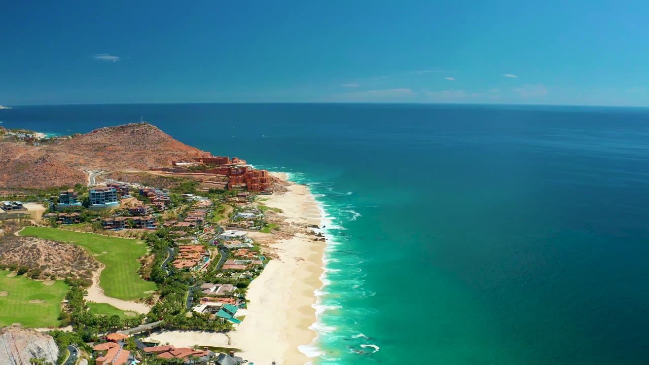 Aerial coastline with white surf edging a sandy beach and buildings below a low hill.