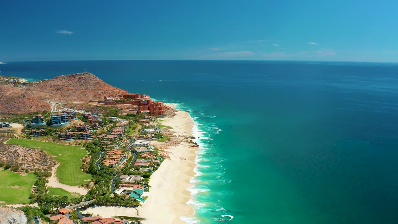 High coastal view with beach, rolling surf, green grounds, buildings, and open ocean.