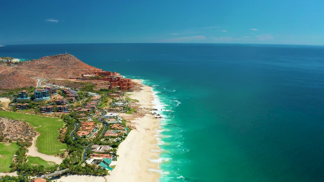 Aerial view of beach, resort buildings, green course, hillside, and turquoise ocean