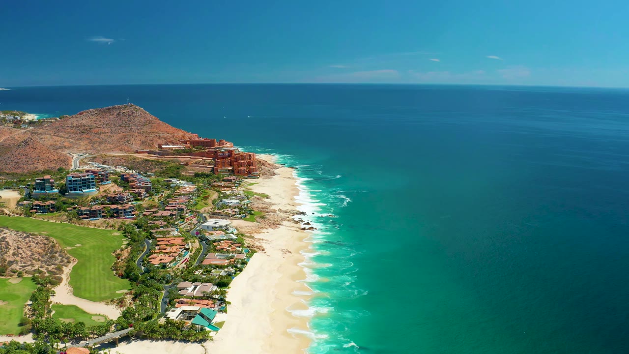 Aerial view of Marquis Los Cabos one second in, with the beach and surf line running diagonally below resort buildings and a wide field of blue ocean, with no visible people.