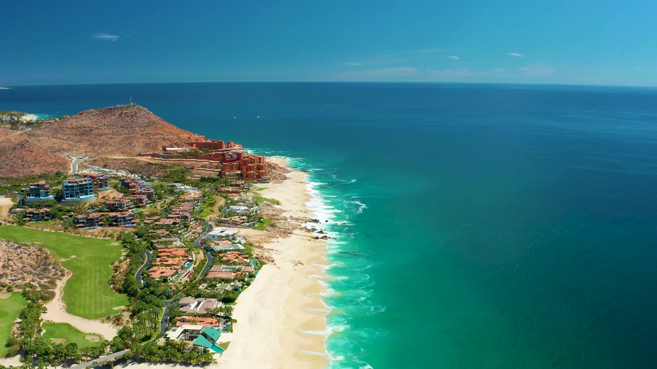 Mid-clip aerial frame of Marquis Los Cabos with a green golf course in the lower left, resort buildings near the beach, white surf along the sand, and no visible people.