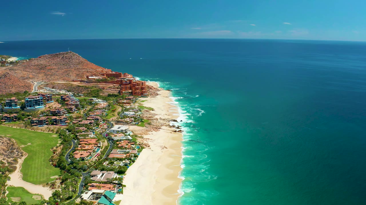 Aerial frame at Marquis Los Cabos, Mexico, showing surf lines edging the sandy beach, resort-area buildings on the hillside, open blue-green ocean, and no visible people.