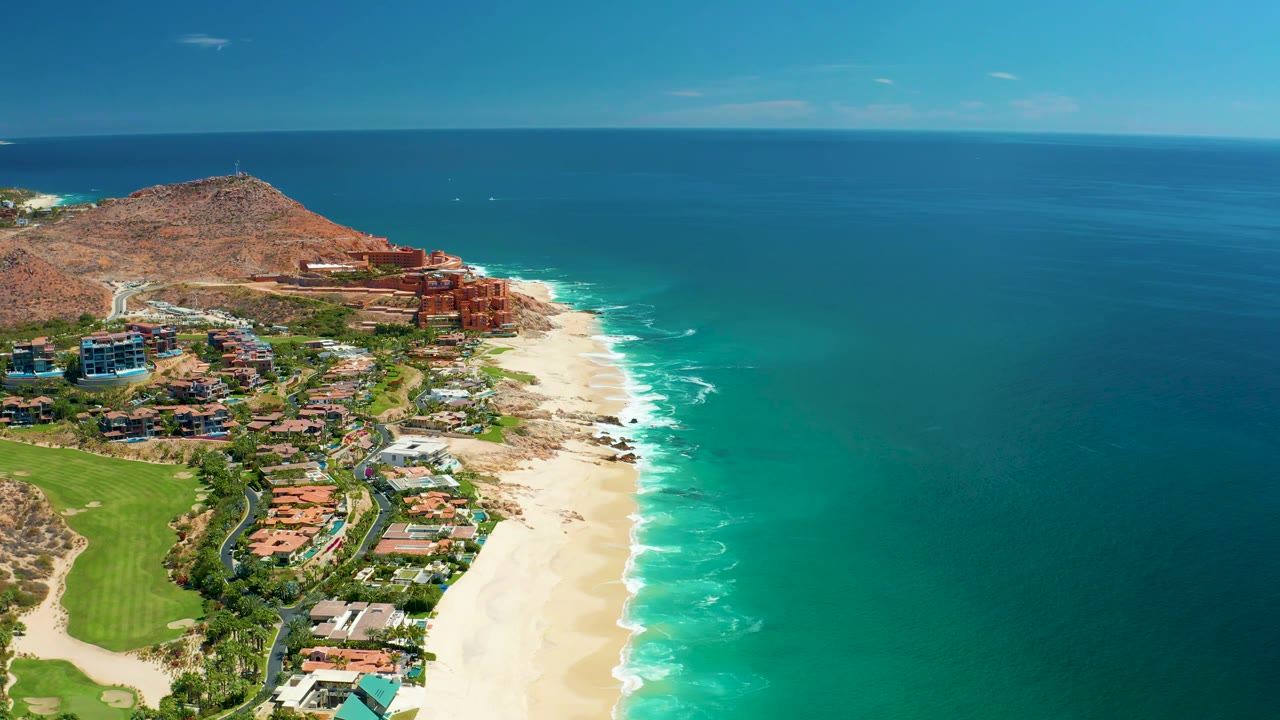 Marquis Los Cabos aerial frame with the beach and hillside buildings held to the left, broad turquoise water filling the right side, thin clouds above, and no visible people.