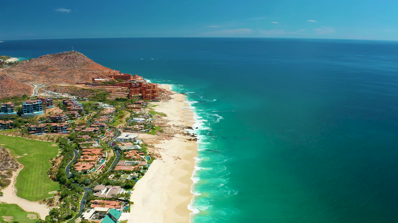 Mid-clip aerial view of Marquis Los Cabos, Mexico, with white waves breaking along the sandy shore, hillside buildings above the beach, green space below, and no visible people.