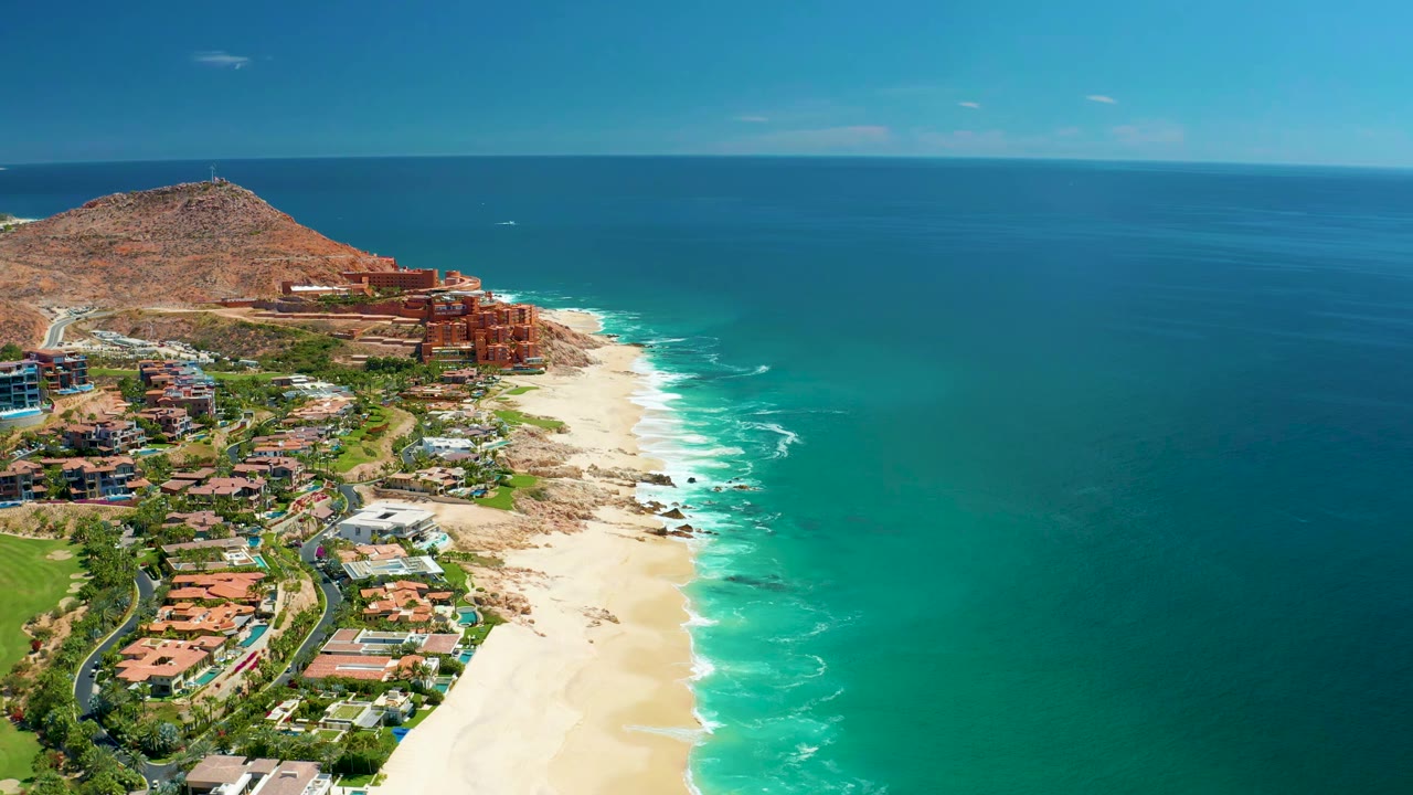 Sandy shoreline with white foam, coastal buildings, hill, and open ocean.