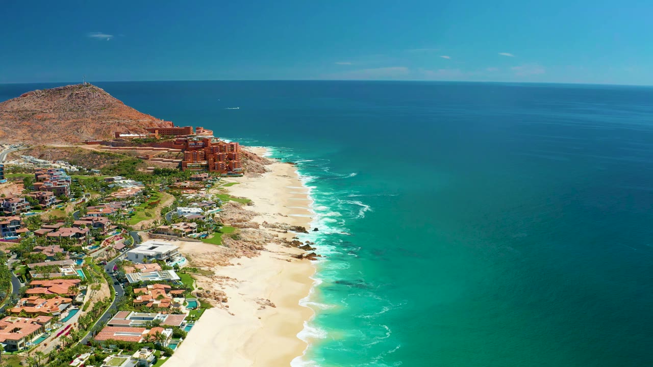 Aerial coastline with waves washing onto sand near coastal buildings.