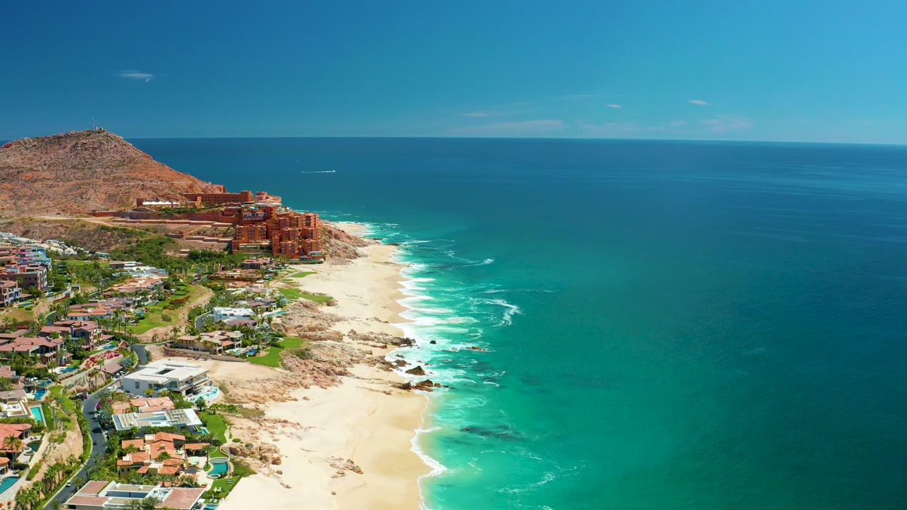 Aerial frame over Marquis Los Cabos, Mexico showing turquoise ocean beside a sandy beach, hillside buildings on the left, a clear blue sky, and no visible people.