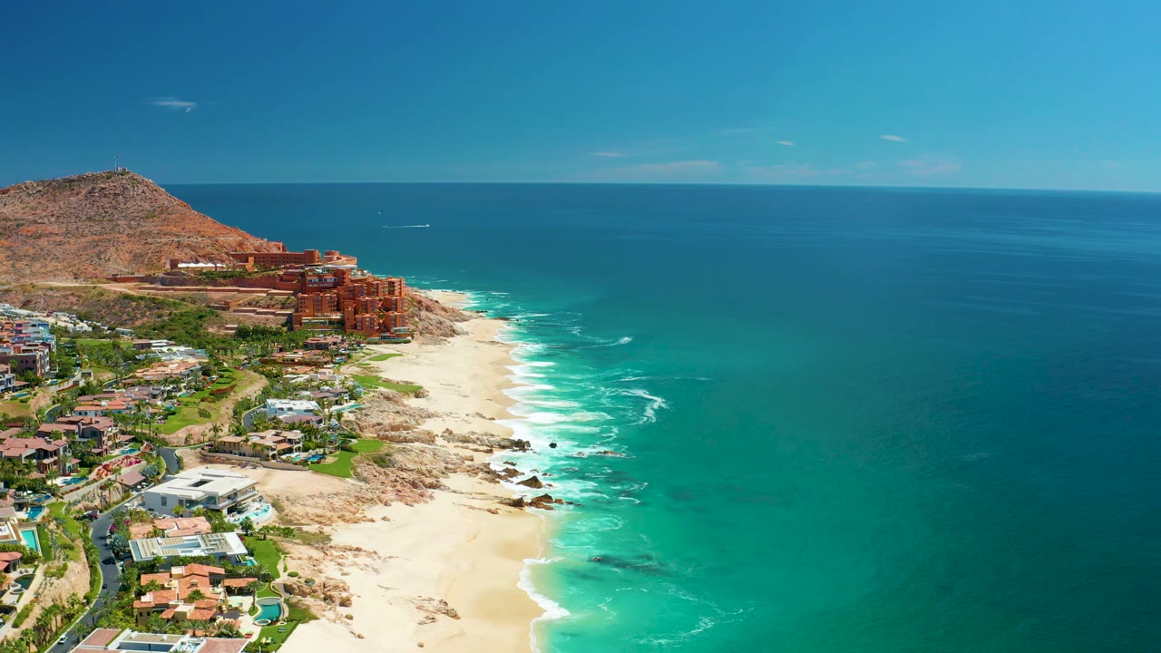 Aerial frame from Marquis Los Cabos, Mexico showing a broad turquoise sea, pale sand, red-toned hillside structures, smaller white buildings, and no visible people.