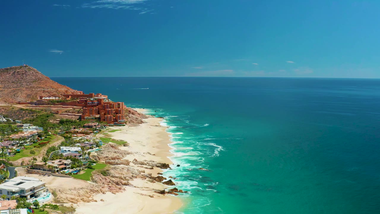 Aerial view at Marquis Los Cabos with turquoise water, white surf, sandy beach, rocky shore, hillside resort buildings, tan mountain, and no visible people.