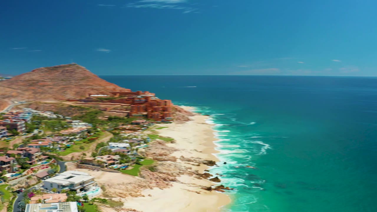 At Marquis Los Cabos, the camera view advances to include more villas and landscaped areas below the hillside resort, with bright surf along the beach and no visible people.