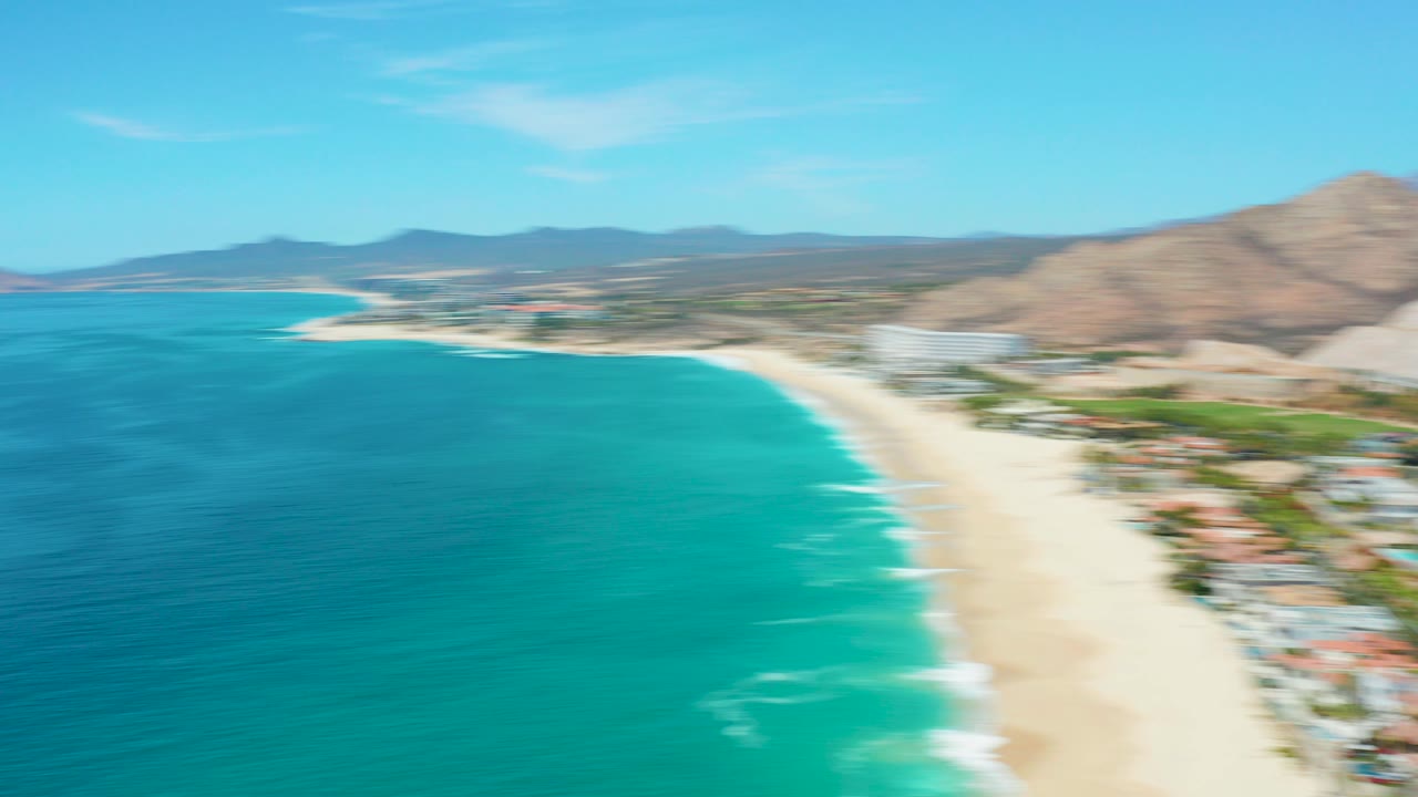 Early aerial frame over Marquis Los Cabos, Mexico with turquoise water, a pale sandy beach, distant coastal buildings, mountains, and no visible people at this scale.