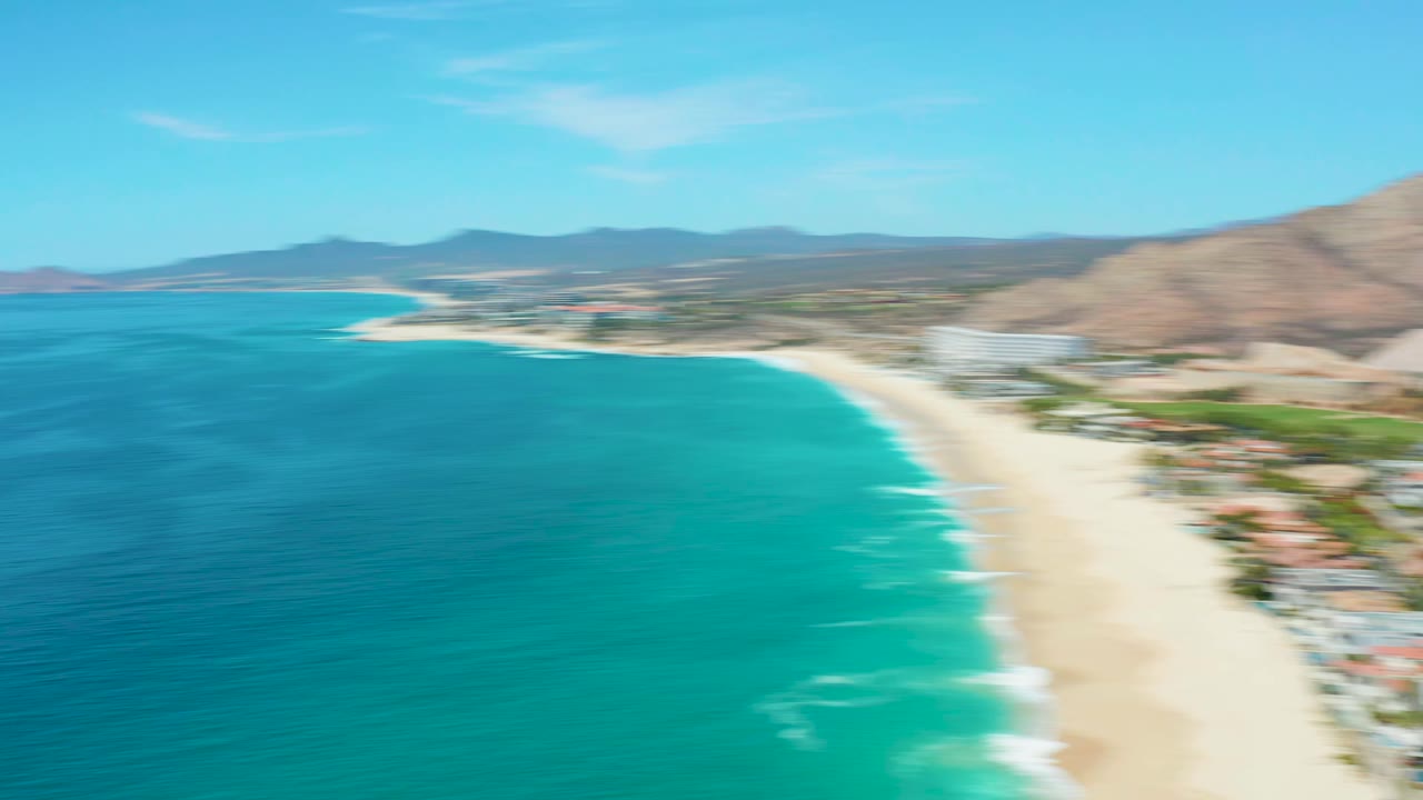 Second aerial frame over Marquis Los Cabos, Mexico showing turquoise ocean on the left, a long sandy beach on the right, resort-area buildings, mountains, and no visible people at this distance.