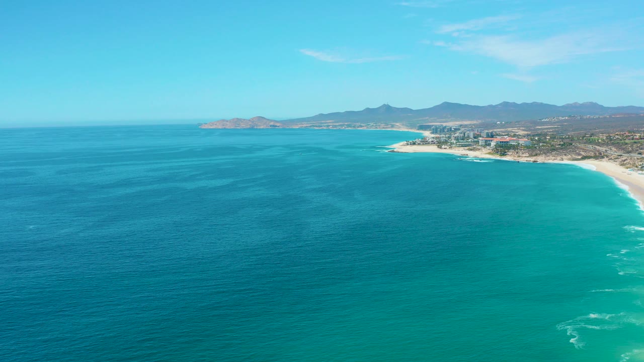 Wide Marquis Los Cabos aerial frame with open turquoise water across the foreground, a curving sandy beach to the right, low coastal development, mountains, and no visible people at this scale.