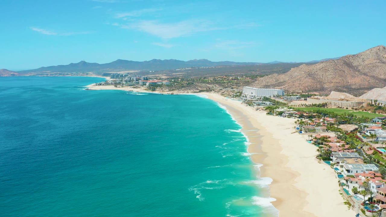 Aerial view along turquoise surf, sandy beach, houses, and mountain slopes.