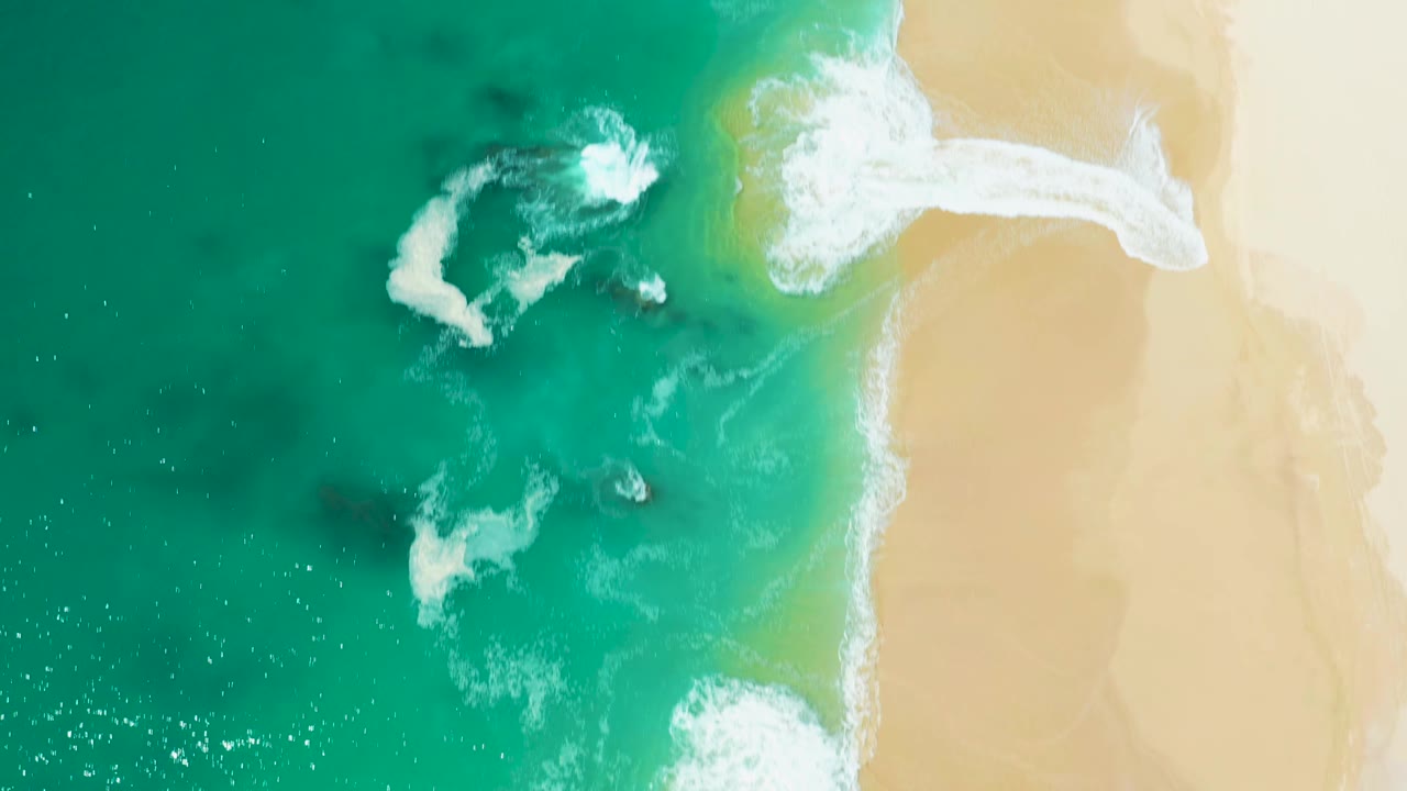 Aerial frame from Marquis Los Cabos, Mexico, showing white foam curling diagonally into turquoise water beside a pale sand beach with no visible people.