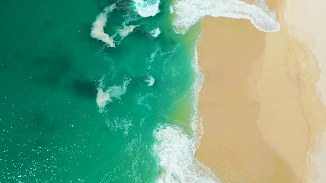 Top-down beach view at Marquis Los Cabos, Mexico, with surf washing from the turquoise ocean onto broad pale sand and no visible people.