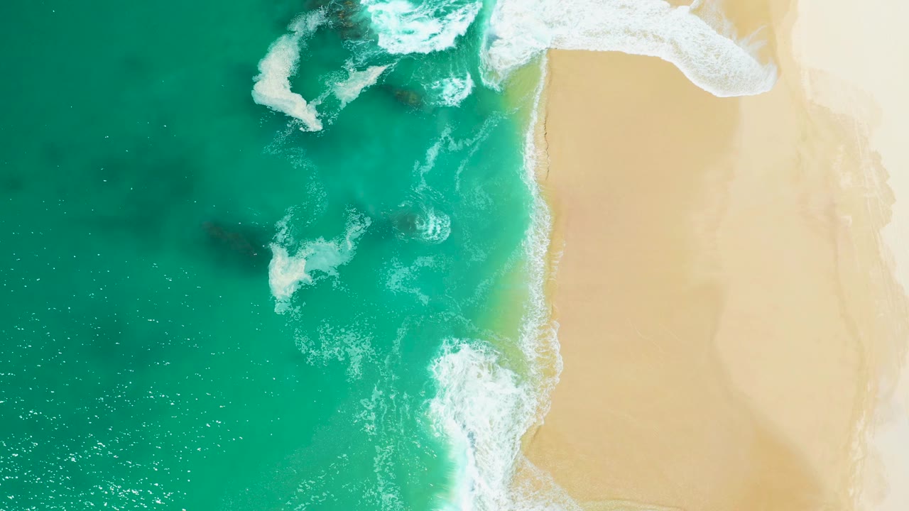 Overhead frame from Marquis Los Cabos, Mexico, with bright white breakers over dark rocks, green-blue water, pale sand on the right, and no visible people.