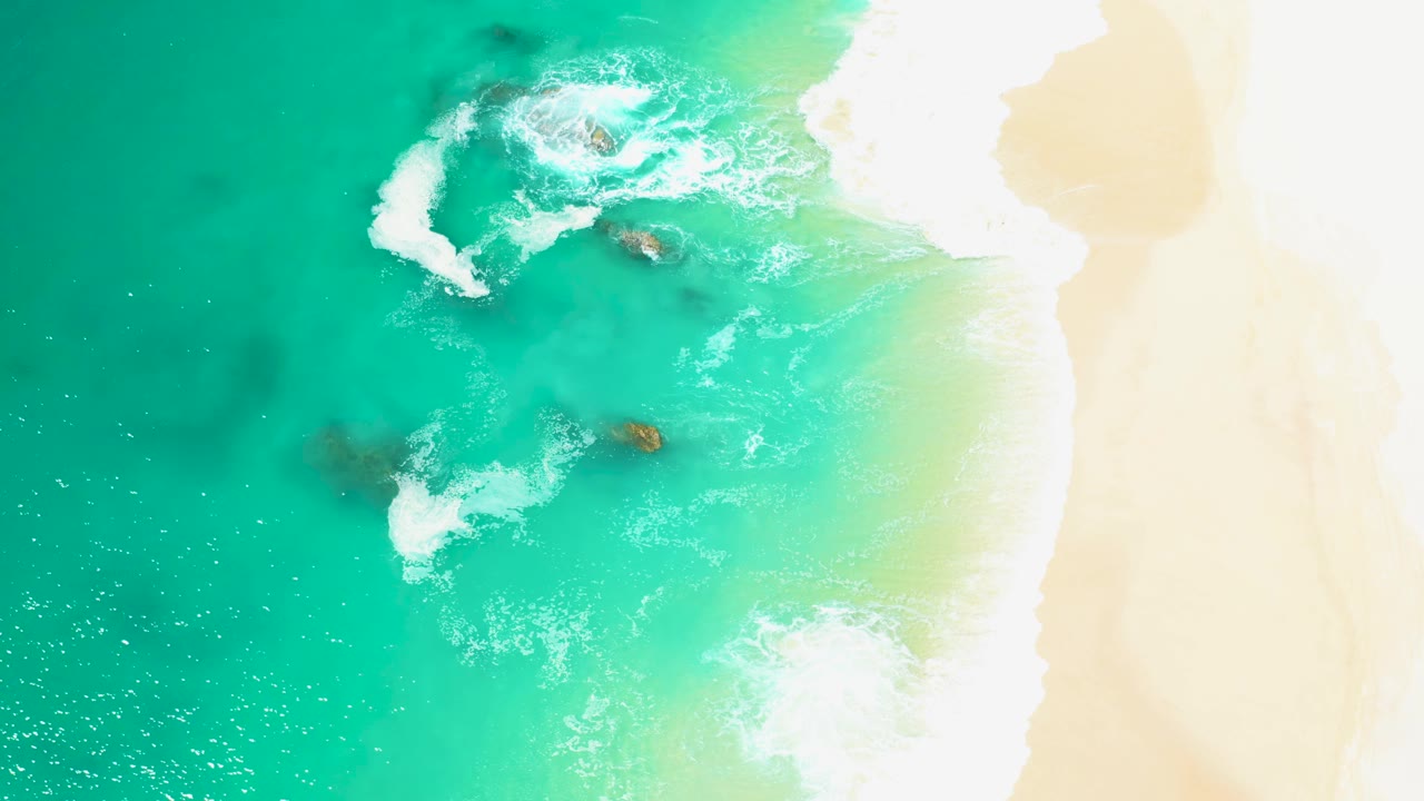 Aerial frame from Marquis Los Cabos with foamy water widening around two central rocks, bright sand along the right edge, and no visible people.