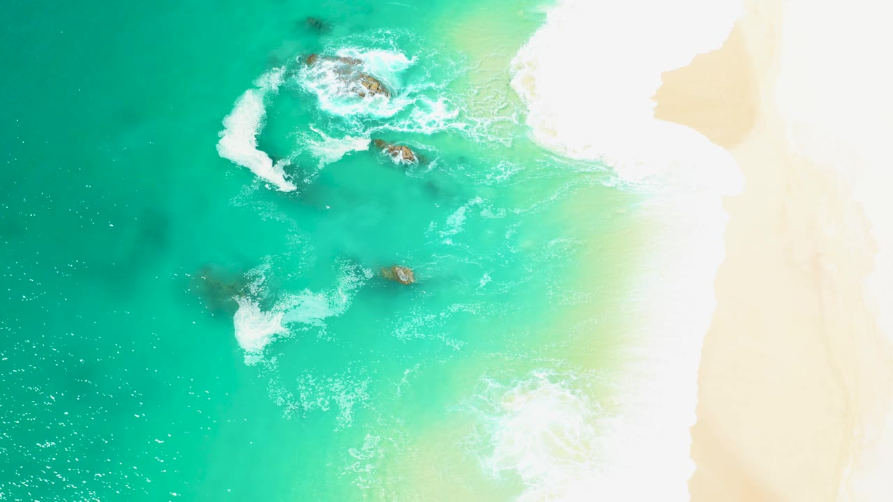 Overhead frame at Marquis Los Cabos where white foam churns around a larger rock cluster near the upper center, smaller rocks sit below, and no visible people appear.
