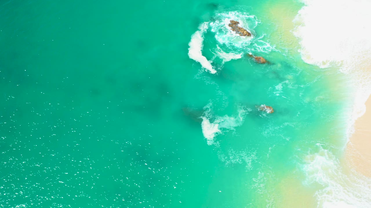 Aerial view at Marquis Los Cabos, Mexico, showing turquoise ocean water, white foam breaking over submerged rocks, pale sand at the right edge, and no visible people.